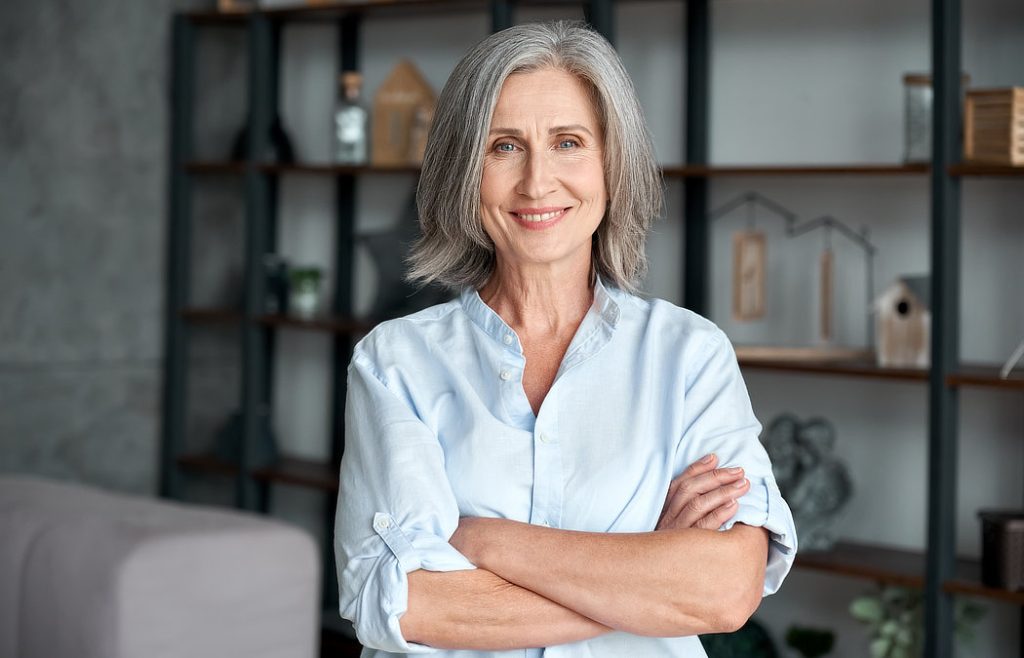 A woman with gray hair and a light blue shirt stands with arms crossed, smiling in a room with shelves in the background.