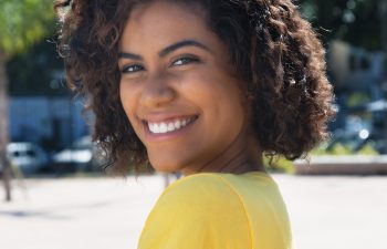 A person with curly hair smiles outdoors while wearing a yellow shirt. Trees and buildings are in the background.