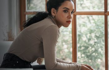 A person with a thoughtful expression sits on a couch by a window, wearing a beige long-sleeve top and dark pants.