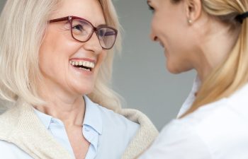 Two women smiling at each other. One has short blonde hair and glasses; the other has long brown hair. They appear to be in a friendly conversation.