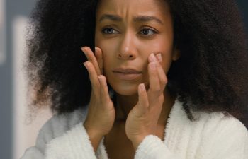 A woman in a white robe touches her face with both hands, looking concerned. She stands indoors with a blurred background.