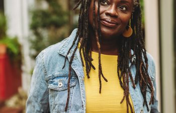 A person with long dreadlocks wearing a denim jacket and a yellow dress stands outdoors.