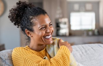 A woman in a yellow sweater smiles while sitting on a couch in a cozy, softly lit living room.