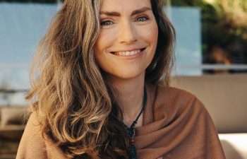 Woman with long hair and a shawl smiles at the camera, with greenery in the background.