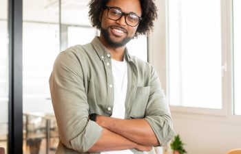 Man with glasses and curly hair stands smiling, arms crossed, in a bright office with a desk and laptop.