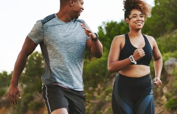 Two people jogging outdoors on a trail, wearing athletic wear and fitness trackers, with greenery in the background.