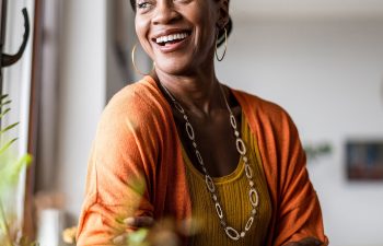 A woman in an orange cardigan smiles while standing near a window. She is wearing a yellow top and a necklace. Plants are visible in the foreground.