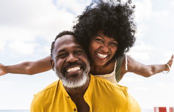 Man in yellow shirt giving a piggyback ride to a joyful woman with curly hair outdoors, both smiling.