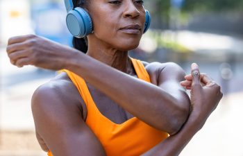 Person wearing headphones and an orange top stretches their arm outdoors.