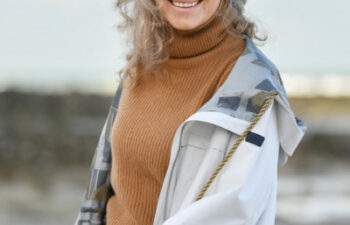Older woman with curly gray hair wearing a tan turtleneck sweater and light jacket, standing outdoors near rocks and water, smiling at the camera.