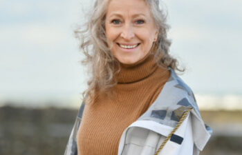 Older woman with curly gray hair wearing a tan turtleneck sweater and light jacket, standing outdoors near rocks and water, smiling at the camera.