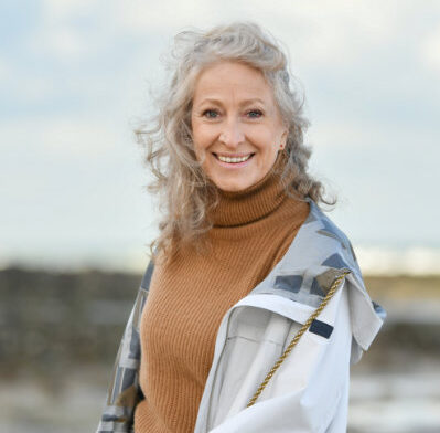 Older woman with curly gray hair wearing a tan turtleneck sweater and light jacket, standing outdoors near rocks and water, smiling at the camera.