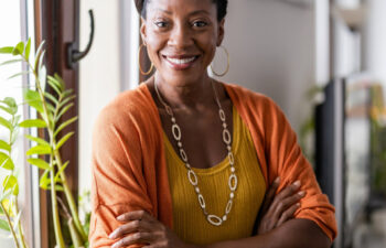 A woman stands indoors by a window, smiling with arms crossed. She wears a mustard top, orange cardigan, hoop earrings, and a necklace. Green plants are visible nearby.