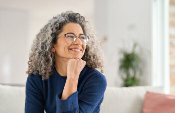 A woman with curly gray hair and glasses sits on a couch, resting her chin on her hand and smiling, with a blurred living room in the background.