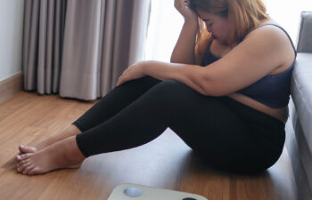 A woman sits on the floor next to a scale, looking down with her hand on her forehead, appearing upset or stressed.