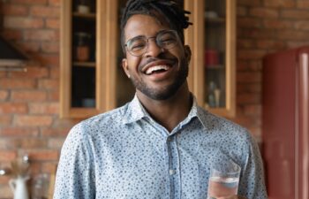 Head shot portrait overjoyed African American man holding glass of fresh pure mineral water, standing in kitchen, looking at camera, healthy lifestyle and good daily habit concept, body and skin care